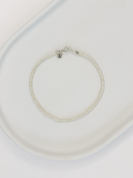 Pulseira de pérolas naturais de 2mm de medida cada, com fecho em prata, apoiada em uma bandeja de cerâmica branca com fundo branco. 