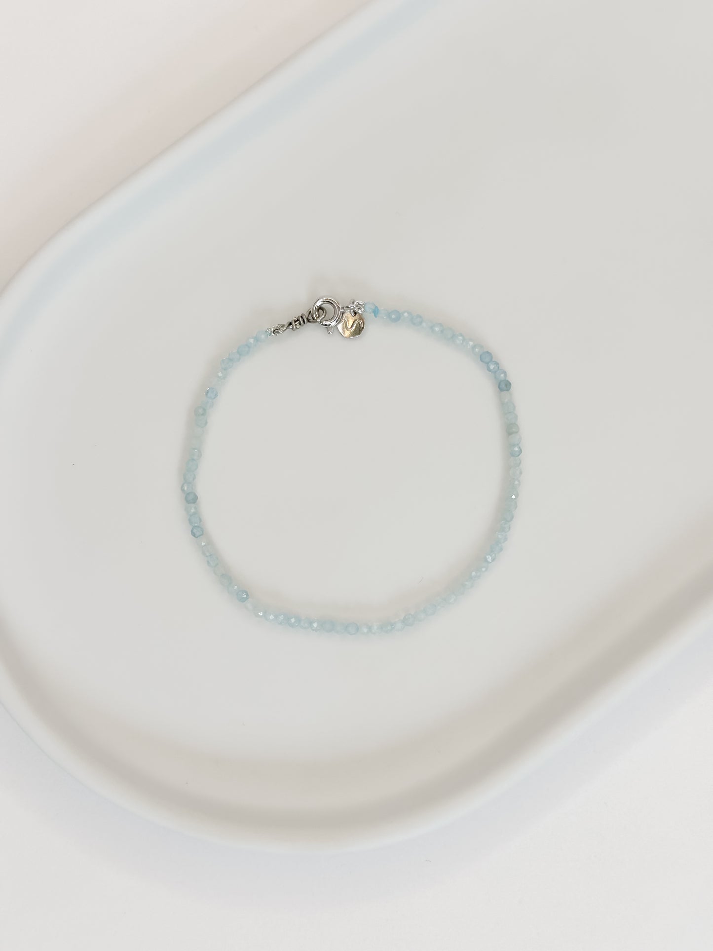 Pulseira ou Tornozeleira produzida com cristais naturais água marinha medindo aproximadamente 2mm cada pedrinha e fecho boia em prata 925 apoiada em ceramica branca com fundo branco.